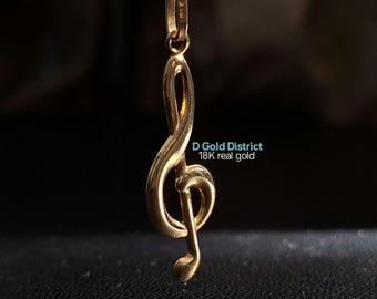 May include: A gold-colored pendant in the shape of a treble clef hangs from a delicate gold chain. The pendant is suspended against a dark background. The text "D Gold District 18K real gold" is visible.
