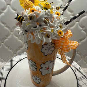 May include: A tall orange mug decorated with white flower outlines, topped with faux white whipped cream, yellow sunflowers, a beehive, and several small bee figures. Two black and white striped straws and an orange gingham bow are attached.