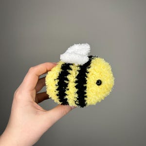 May include: A crocheted bee toy with yellow and black stripes and a white fuzzy head. The bee has a black button eye.