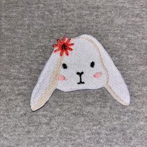 May include: Embroidered white bunny face with long ears, black eyes, and a black nose. The bunny has pink cheeks and a red flower on its head. The bunny is on a gray background.