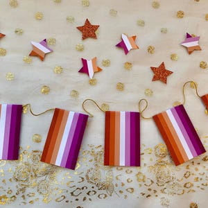 Puede incluir: Una pancarta con rayas alternas de color naranja, blanco y morado, enhebrada con hilo dorado y decorada con estrellas de purpurina.