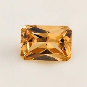 Puede incluir: Una gema de color amarillo dorado, de corte rectangular, con múltiples facetas. La gema se encuentra sobre un fondo blanco liso, mostrando su claridad y brillo. El corte de la piedra crea un juego de luces y sombras, resaltando su color y forma.