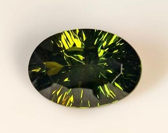 May include: An oval-cut gemstone with a dark green hue and vibrant yellow internal reflections. The gem's facets catch the light, creating a striking contrast between the dark base color and the bright yellow flashes. The gemstone is set against a neutral background.