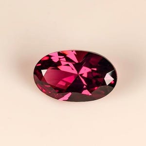 Puede incluir: Una gema ovalada y facetada de color magenta intenso. La gema tiene múltiples facetas que reflejan la luz. La piedra preciosa se muestra sobre un fondo neutro, resaltando su color vibrante y su talla.