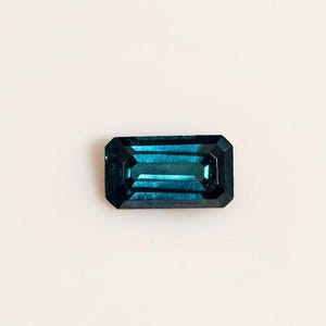 以下が含まれることがあります： 深いティールブルーの色をした、長方形のエメラルドカットの宝石。石にはファセットカットされたエッジと光を反射する研磨された表面があり、幾何学的なラインがはっきりと見える透明な内部構造を特徴としています。
