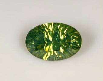 May include: An oval-shaped, faceted gemstone with a vibrant green hue and yellow highlights. The gemstone's cut creates a play of light, enhancing its brilliance. The gem is set against a plain white background.
