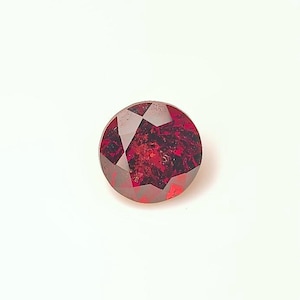 May include: A faceted, round, deep red gemstone. The gem has a complex internal structure with dark inclusions, and is set against a white background. The cut of the stone creates multiple reflective surfaces.