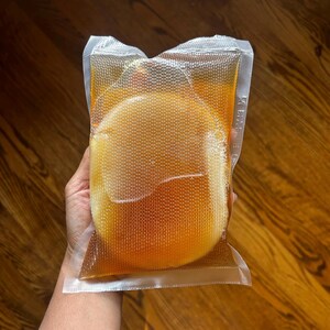 Op de afbeelding: Een doorzichtige, verzegelde plastic zak met een goudbruine vloeistof en een rond, lichtgekleurd object. De zak heeft een gestructureerd oppervlak en wordt tegen een houten achtergrond gehouden. De vloeistof lijkt een drankje te zijn.