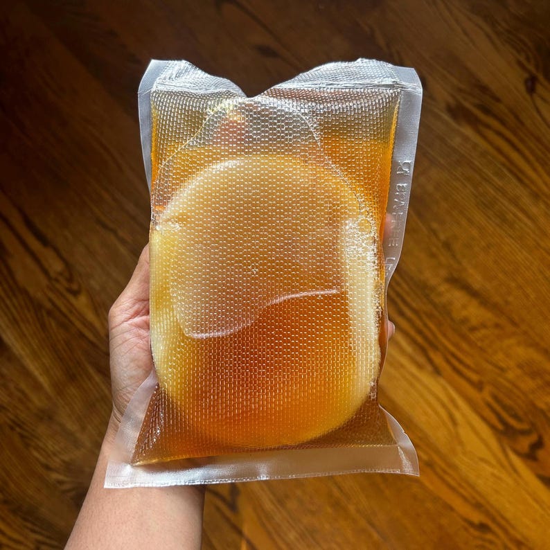 Op de afbeelding: Een doorzichtige, verzegelde plastic zak met een goudbruine vloeistof en een rond, lichtgekleurd object. De zak heeft een gestructureerd oppervlak en wordt tegen een houten achtergrond gehouden. De vloeistof lijkt een drankje te zijn.