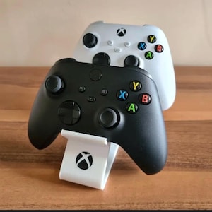 May include: Two wireless game controllers, one black and one white, are stacked on a wooden surface. The black controller rests on a white stand with the Xbox logo. The white controller is behind the black one. Both controllers have colored buttons.