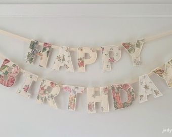 Peut inclure: Une banderole en tissu à deux rangées épelant HAPPY BIRTHDAY en lettres capitales. Chaque lettre est fabriquée dans un tissu crème avec un motif floral multicolore dans des tons de rose, vert et jaune, suspendue sur un ruban crème uni.