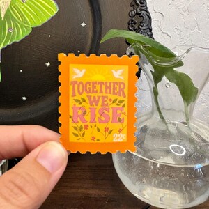May include: An orange postage stamp with the words "TOGETHER WE RISE" in pink and yellow, with floral and bird accents. The stamp has a 22&cent; value and is held in front of a black plate with a green moth illustration.