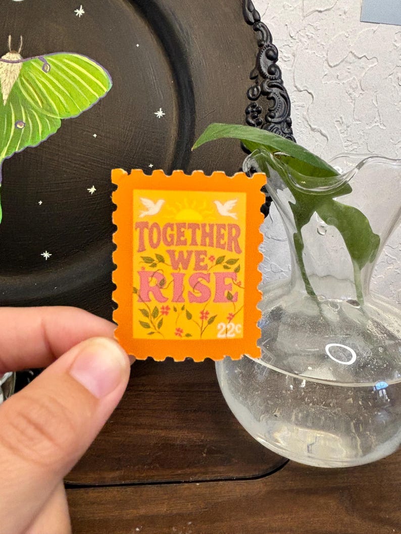 May include: An orange postage stamp with the words "TOGETHER WE RISE" in pink and yellow, with floral and bird accents. The stamp has a 22&cent; value and is held in front of a black plate with a green moth illustration.