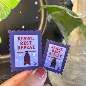 May include: Two purple postage stamp stickers with the words "RESIST. REST. REPEAT." A brown bat hangs upside down from a branch, with a crescent moon and stars. The stickers are 5&cent; each.