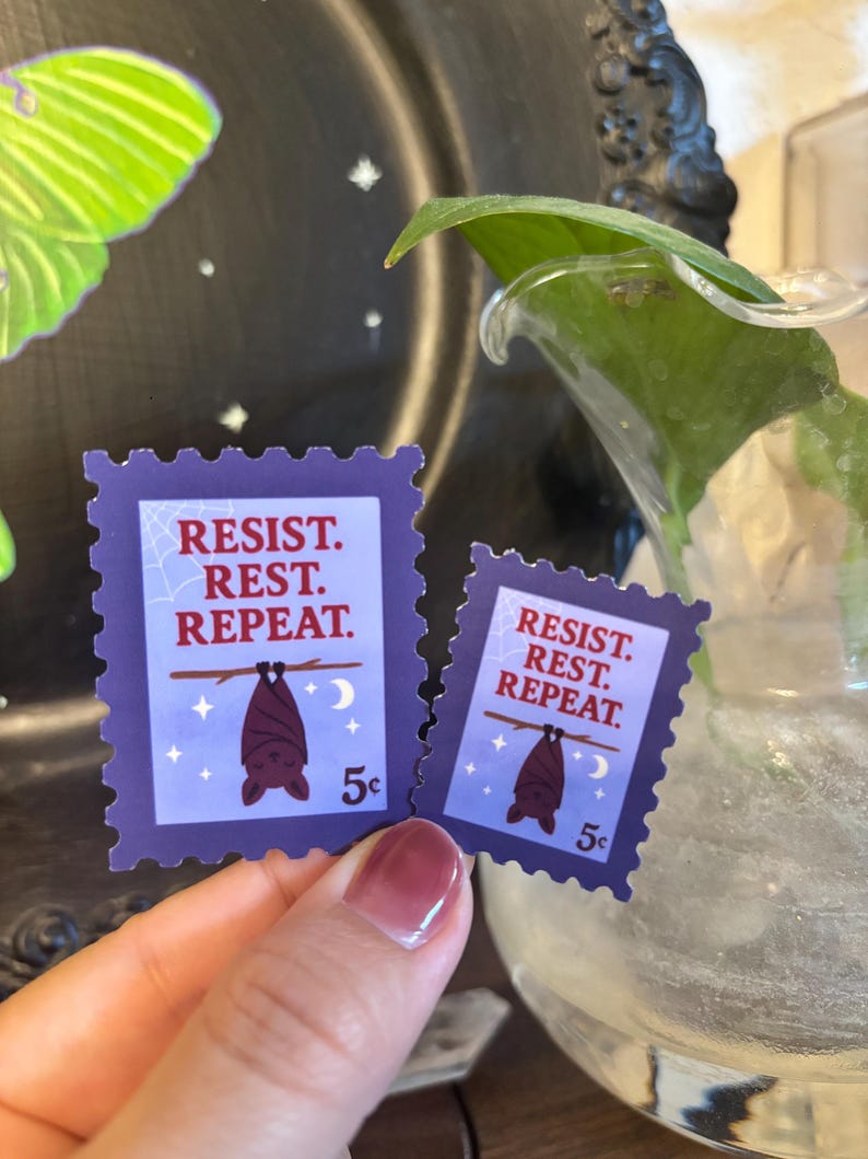 May include: Two purple postage stamp stickers with the words "RESIST. REST. REPEAT." A brown bat hangs upside down from a branch, with a crescent moon and stars. The stickers are 5&cent; each.
