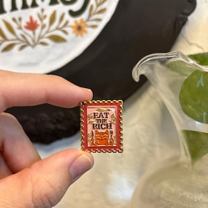 May include: A rectangular enamel pin with a red border and gold trim. The pin features a pink background with the words "EAT THE RICH" and an orange cat holding a knife and fork. The pin is held by a person.