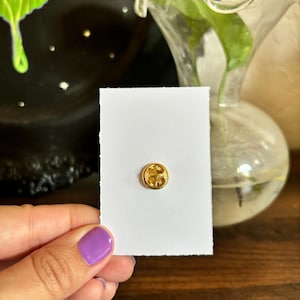 May include: A small, round, gold-colored pin with a decorative design is affixed to a white card with a torn edge. The pin is the focal point, held by a person with purple nail polish. A black decorative plate and a glass vase are in the background.