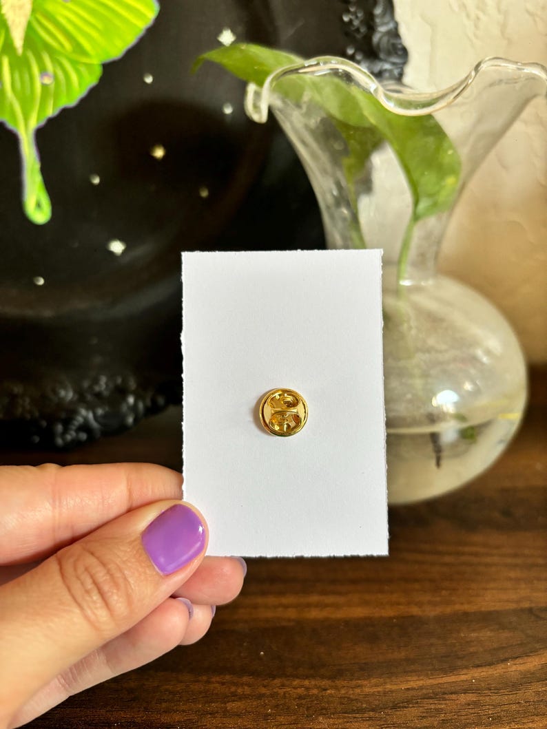 May include: A small, round, gold-colored pin with a decorative design is affixed to a white card with a torn edge. The pin is the focal point, held by a person with purple nail polish. A black decorative plate and a glass vase are in the background.