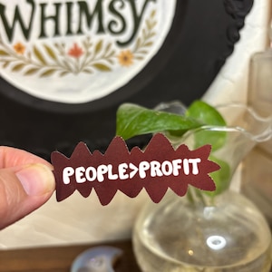 May include: A maroon-colored sticker with the text "PEOPLE>PROFIT" in white, held in a hand. The sticker has a jagged edge design. In the background, a decorative frame with the words "RAGE & WHIMSY" is visible.
