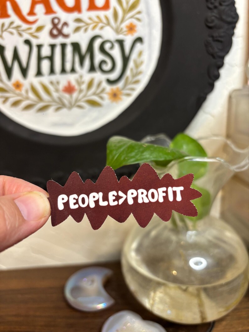 May include: A maroon-colored sticker with the text "PEOPLE>PROFIT" in white, held in a hand. The sticker has a jagged edge design. In the background, a decorative frame with the words "RAGE & WHIMSY" is visible.