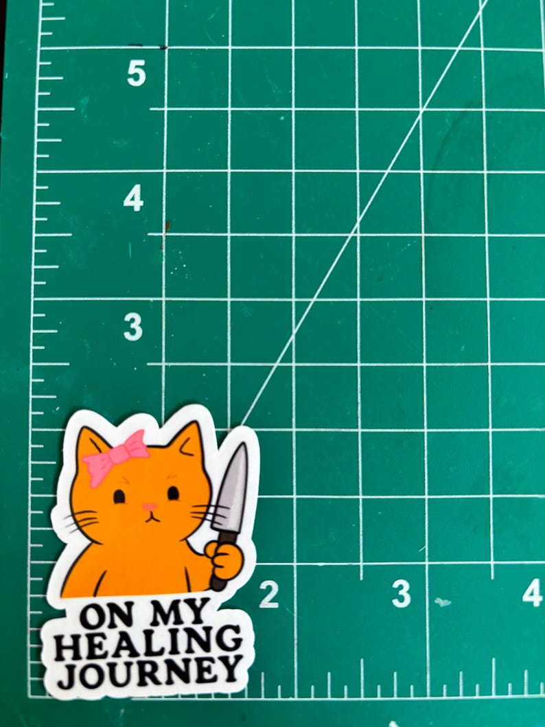 May include: Orange cat sticker with a pink bow holding a knife. The sticker has the text "ON MY HEALING JOURNEY" in a black font. The sticker is on a green cutting mat.