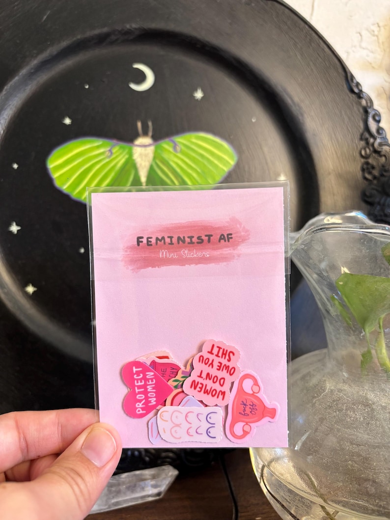 May include: A pink packet of stickers with the text "FEMINIST AF" and "Mira Stickers". The stickers have various designs and phrases, including "PROTECT WOMEN" and "WOMEN DON'T OWE YOU SHIT". A black plate with a green moth and moon is in the background.