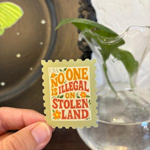 May include: A rectangular sticker resembling a postage stamp, held by a hand. The sticker has a light green background with the words "NO ONE IS ILLEGAL ON STOLEN LAND" in orange and red. Small orange flowers are also present.
