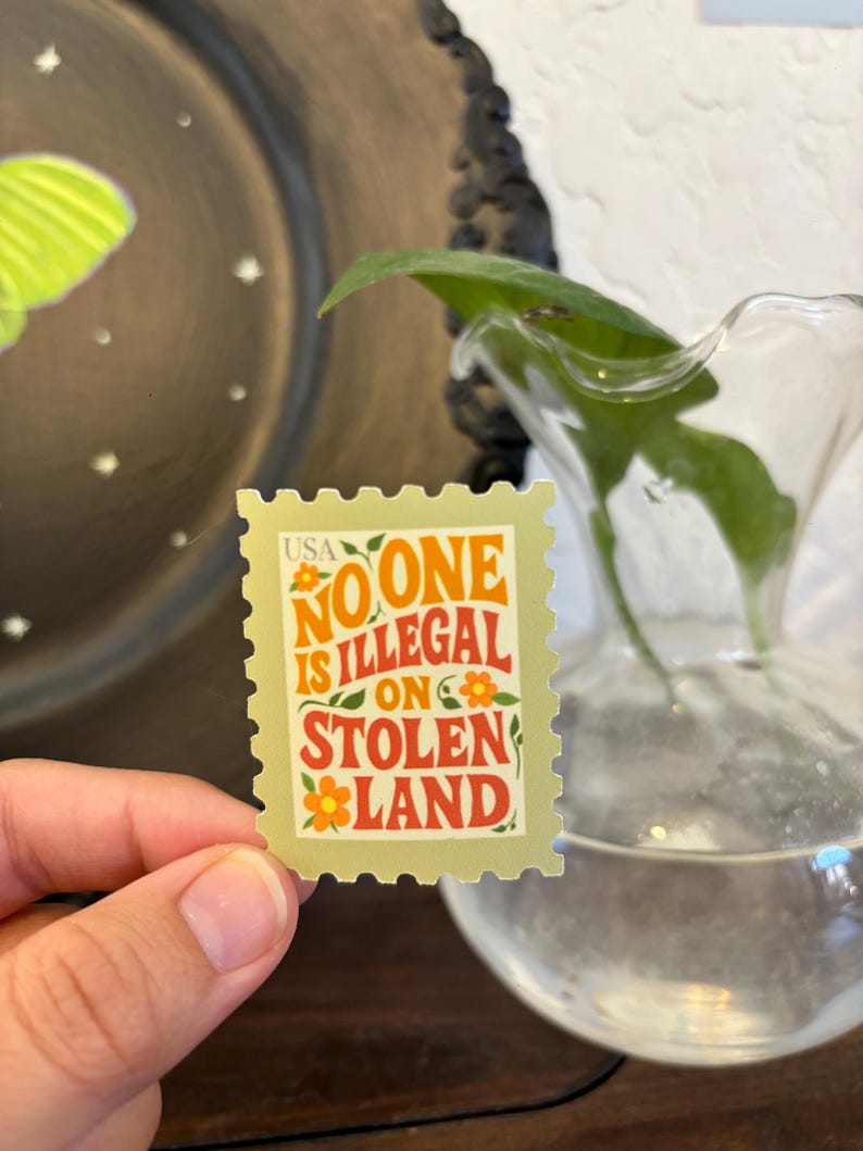 May include: A rectangular sticker resembling a postage stamp, held by a hand. The sticker has a light green background with the words "NO ONE IS ILLEGAL ON STOLEN LAND" in orange and red. Small orange flowers are also present.