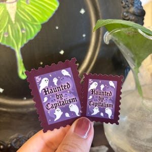 May include: Two purple postage stamps with a dark burgundy border. The stamps feature white ghost illustrations and the text "Haunted by Capitalism" in a stylized font. The stamps are held in front of a black background.