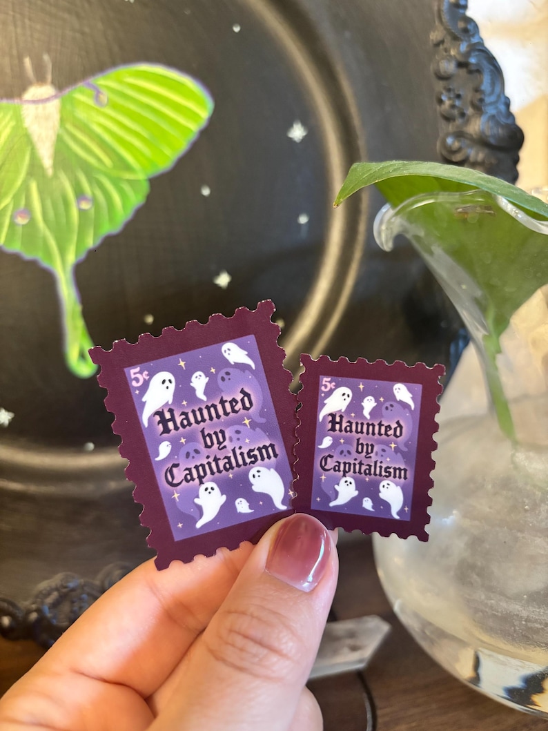 May include: Two purple postage stamps with a dark burgundy border. The stamps feature white ghost illustrations and the text "Haunted by Capitalism" in a stylized font. The stamps are held in front of a black background.