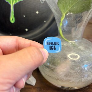 May include: A small, blue sticker with the words "ABOLISH ICE" in white block letters. The sticker is held in a person's hand, with a blurred background featuring a glass vase and a dark, painted surface.