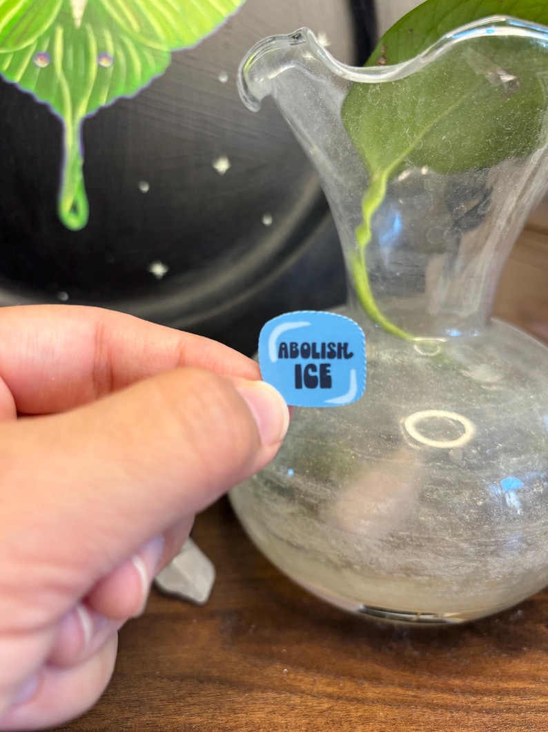 May include: A small, blue sticker with the words "ABOLISH ICE" in white block letters. The sticker is held in a person's hand, with a blurred background featuring a glass vase and a dark, painted surface.