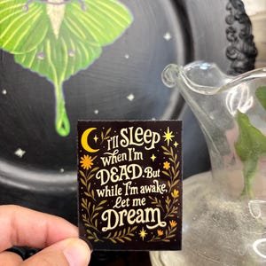 May include: A rectangular black sticker with the text "I'll Sleep when I'm DEAD. But while I'm awake, Let me Dream" in white and gold lettering. The sticker has floral and celestial accents. A green moth is in the background.