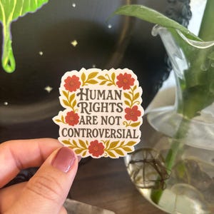 May include: A sticker with the text "HUMAN RIGHTS ARE NOT CONTROVERSIAL" in a floral design with red flowers and gold leaves. The sticker is held up in front of a blurred background.