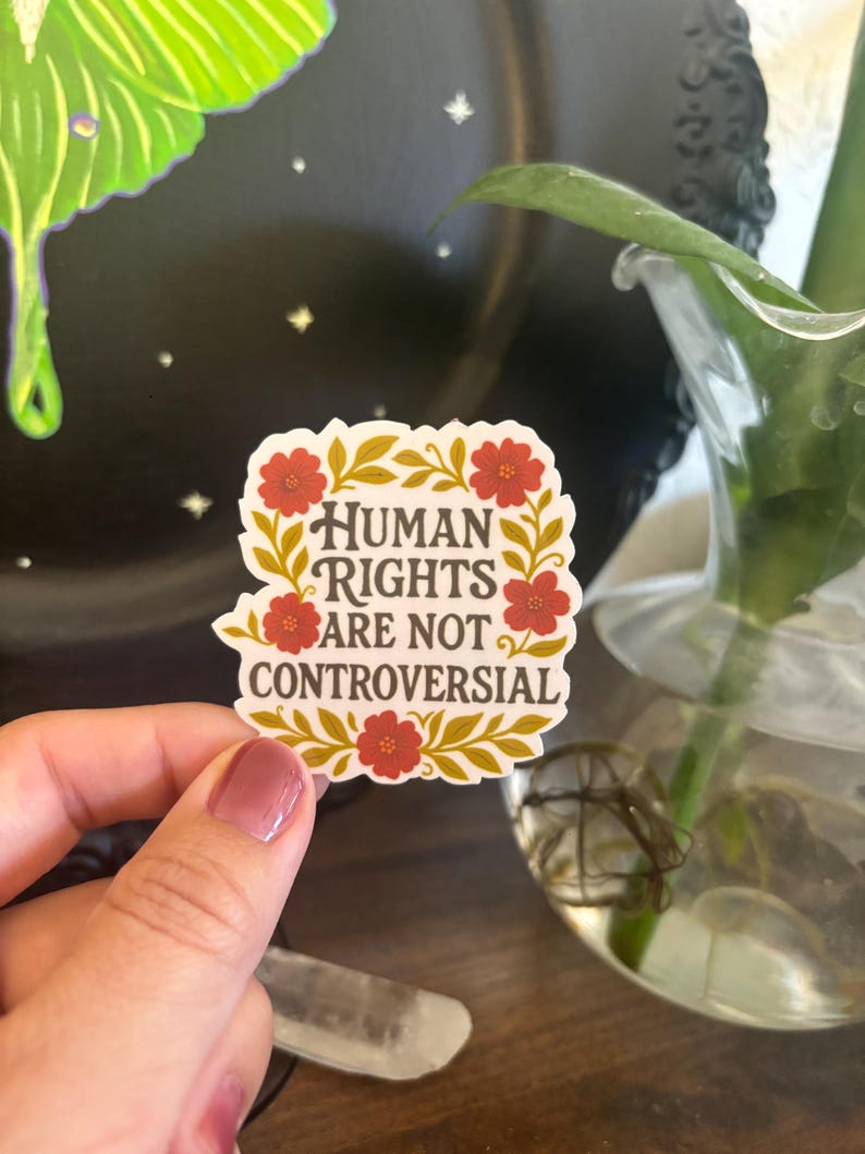 May include: A sticker with the text "HUMAN RIGHTS ARE NOT CONTROVERSIAL" in a floral design with red flowers and gold leaves. The sticker is held up in front of a blurred background.
