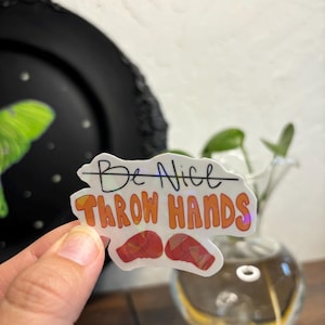 Può includere: Un adesivo bianco con il testo "Be Nice" barrato e il testo "Throw Hands" in arancione. L'adesivo ha una finitura olografica e presenta due guantoni da boxe rossi.