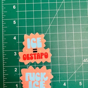 Puede incluir: Dos pegatinas con fondo melocot&oacute;n y texto azul y rojo. Una pegatina dice "ICE = GESTAPO" y la otra "FUCK ICE". Las pegatinas est&aacute;n sobre una esterilla de corte verde con medidas en pulgadas, equivalentes a 2,54 cm.
