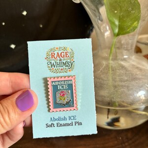May include: A soft enamel pin with a pink border and the words "Abolish ICE" on a light blue card. The pin features a postage stamp design with a floral motif. The card also has the text "Rage & Whimsy".