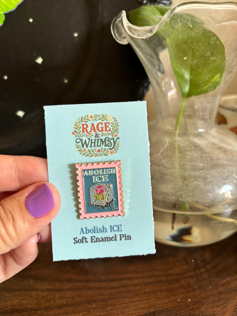May include: A soft enamel pin with a pink border and the words "Abolish ICE" on a light blue card. The pin features a postage stamp design with a floral motif. The card also has the text "Rage & Whimsy".