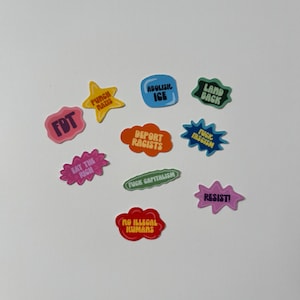 May include: A collection of colorful stickers with various phrases and slogans. The stickers are in different shapes, including stars, speech bubbles, and ovals. The text includes phrases like "Abolish ICE," "Resist!," and "Eat the Rich."