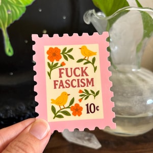 Könnte beinhalten: Ein rosa Aufkleber in Form einer Briefmarke mit den Worten "FUCK FASCISM" in brauner Schrift. Der Aufkleber zeigt gelbe Vögel, orangefarbene Blumen und die Zahl "10¢".