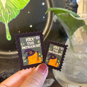 May include: Two postage stamp-style stickers with a dark purple border. Each sticker features an orange cat wearing a witch hat, holding a magic wand, and the text "HEX THE PATRIARCHY". The stickers are 10&cent; each.