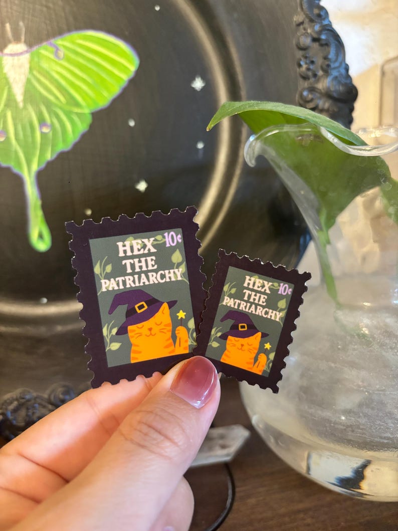 May include: Two postage stamp-style stickers with a dark purple border. Each sticker features an orange cat wearing a witch hat, holding a magic wand, and the text "HEX THE PATRIARCHY". The stickers are 10&cent; each.