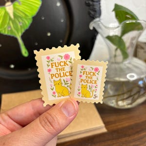 a stamp shaped sticker featuring a cute cat surrounded by flowers and leaves. the text reads “fuck the police.”