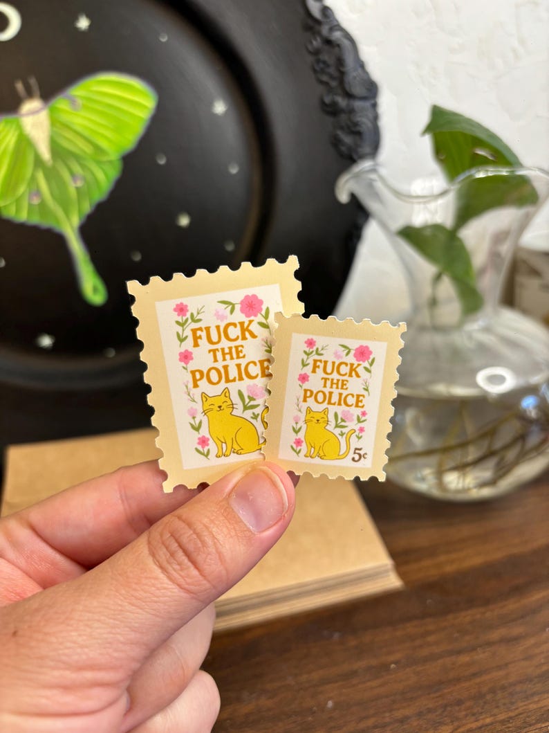 a stamp shaped sticker featuring a cute cat surrounded by flowers and leaves. the text reads “fuck the police.”