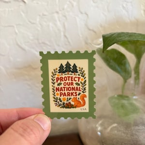 K&ouml;nnte beinhalten: Ein gr&uuml;ner und cremefarbener Aufkleber in Form einer Briefmarke. Der Aufkleber zeigt den Text "PROTECT OUR NATIONAL PARKS" mit einem Eichh&ouml;rnchen und B&auml;umen. Das Wort "USA" steht unten.