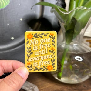 以下が含まれることがあります： 「No one is free until everyone is free」のテキストが入った黄色のステッカー。オレンジ色の花と緑の葉のフローラルボーダーがあります。ステッカーは手で持たれています。