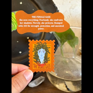 May include: An orange stamp with the text "THE FEMALE GAZE" and a Medusa illustration. The stamp is held in a hand, with a clear glass vase and a decorative orange sign in the background. The sign reads "She sees everything. Fearlessly, she confronts her shadows."