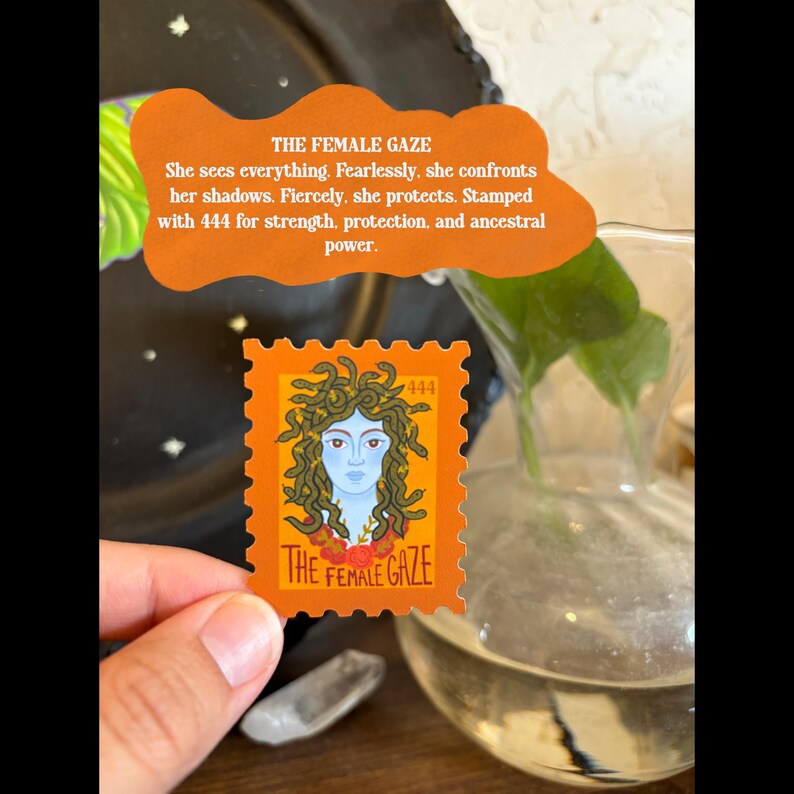May include: An orange stamp with the text "THE FEMALE GAZE" and a Medusa illustration. The stamp is held in a hand, with a clear glass vase and a decorative orange sign in the background. The sign reads "She sees everything. Fearlessly, she confronts her shadows."