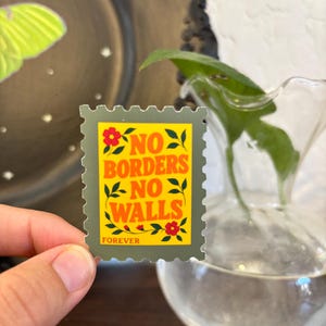 May include: A green postage stamp with the words "NO BORDERS NO WALLS" in orange lettering. The stamp features red flowers and green leaves. The word "FOREVER" is printed at the bottom. The stamp is held in a person's hand.